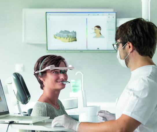 Zahnärztliche Untersuchung mit digitalem Gesichtsbogen zur Erfassung von Kieferbewegungen, auf Monitoren sind 3D-Modelle des Gebisses sichtbar.