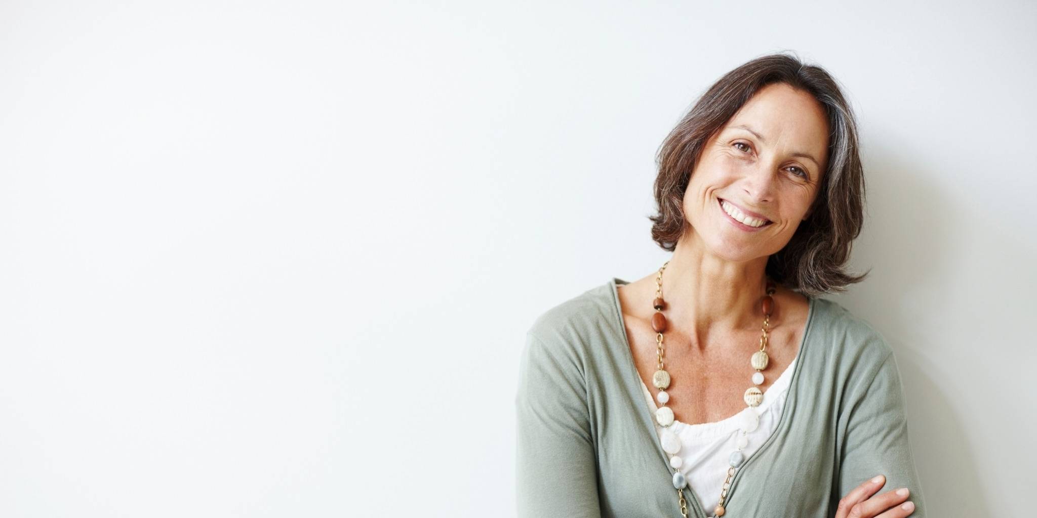 Reifere Frau mit halblangen braunen Haaren steht lächelnd mit verschränkten Armen vor einer weißen Wand, trägt eine lange Kette und einen hellgrünen Cardigan.
