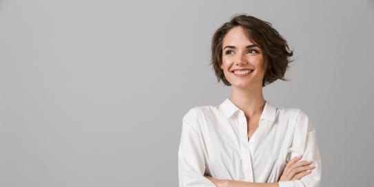 Junge Frau mit kurzem braunen Haar lächelt und verschränkt die Arme, vor grauem Hintergrund.