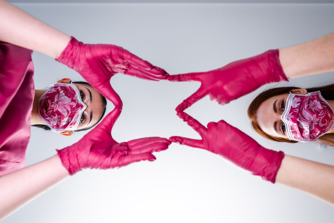 Zwei Frauen mit gemusterten Masken und pinken Handschuhen formen gemeinsam mit ihren Händen ein Herzsymbol in Richtung Kamera.