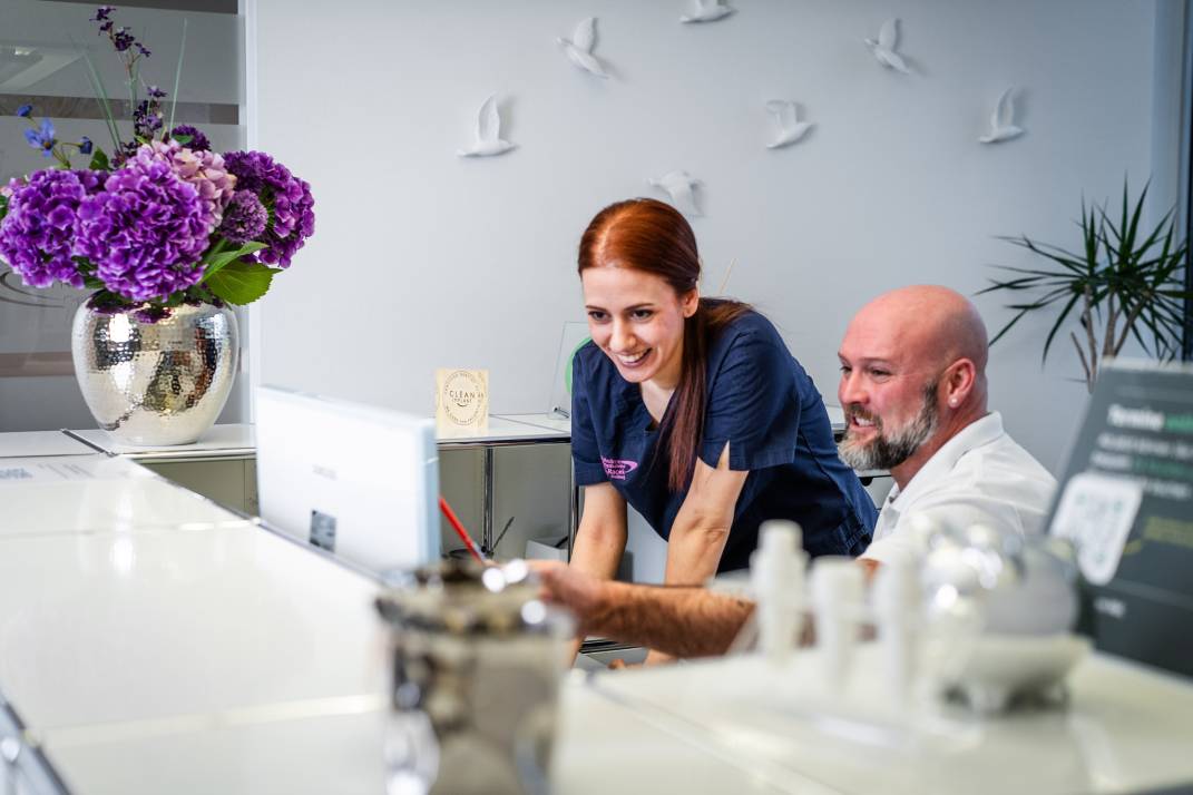 Mitarbeiterin in medizinischer Kleidung und ein Patient schauen gemeinsam auf einen Bildschirm an der Rezeption einer modernen Zahnarztpraxis.