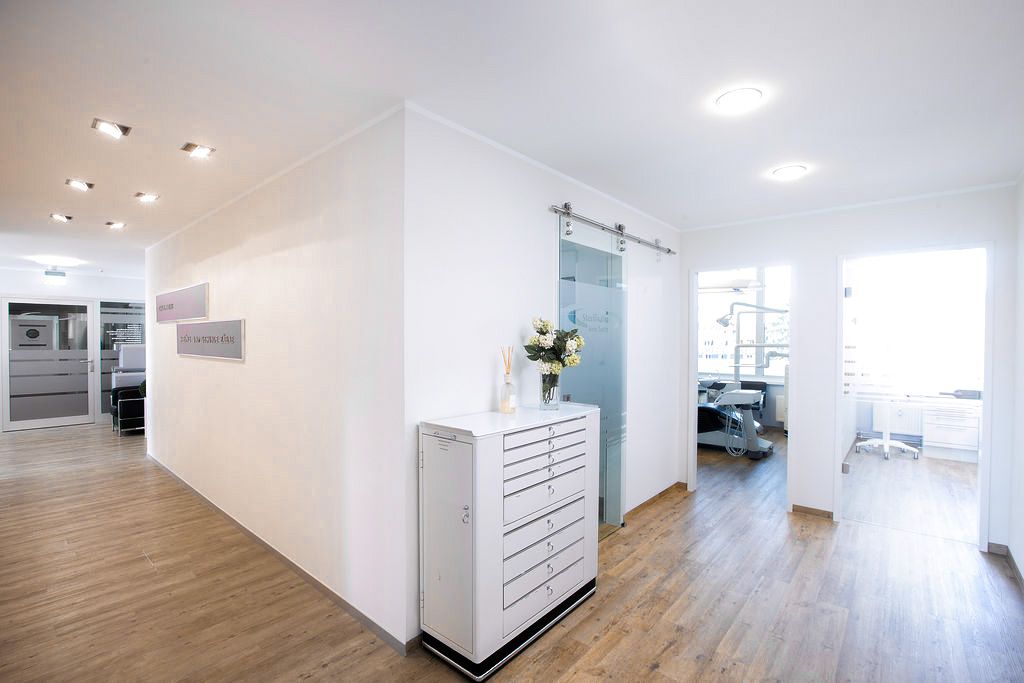 Heller Flur einer Zahnarztpraxis mit Holzfußboden, modernen Glastüren und Blick in zwei Behandlungsräume.