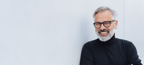 Älterer Mann mit grauem Haar, Bart und Brille steht lächelnd mit verschränkten Armen vor einer hellen Wand, trägt einen schwarzen Rollkragenpullover.