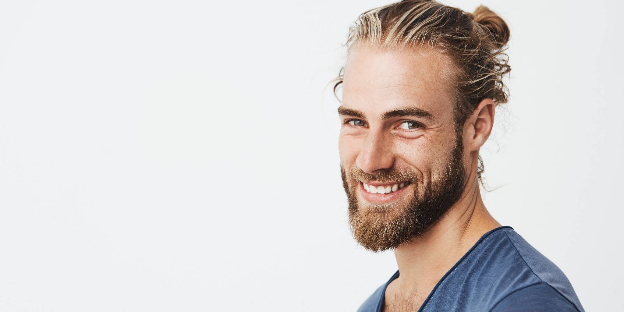 Junger Mann mit Bart und zurückgebundenen Haaren lächelt in die Kamera vor hellem Hintergrund.