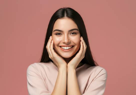 Junge Frau mit langen dunklen Haaren lächelt und hält ihr Gesicht mit beiden Händen, vor rosa Hintergrund.