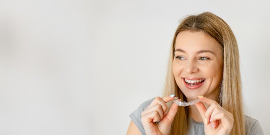 Junge Frau mit langen blonden Haaren lächelt und hält eine transparente Zahnschiene in den Händen.