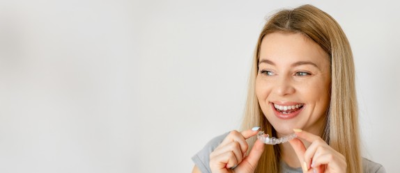 Junge Frau mit langen blonden Haaren lächelt und hält eine transparente Zahnschiene in den Händen.