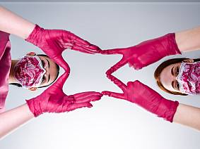 Zwei Frauen mit gemusterten Masken und pinken Handschuhen formen gemeinsam mit ihren Händen ein Herzsymbol in Richtung Kamera.