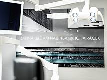 Wandbild mit moderner Architektur und dem Schriftzug „Zahnärzte am Hauptbahnhof // Racek – seit 2013“ in einer Zahnarztpraxis, im Vordergrund ein Teil der Behandlungseinheit.