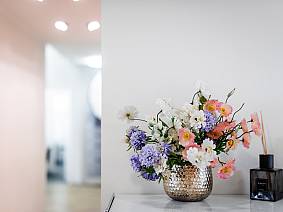 Bunter Blumenstrauß in einer strukturierten silbernen Vase auf einem weißen Sideboard, daneben ein Raumduftspender in einem dunklen Glasflakon.